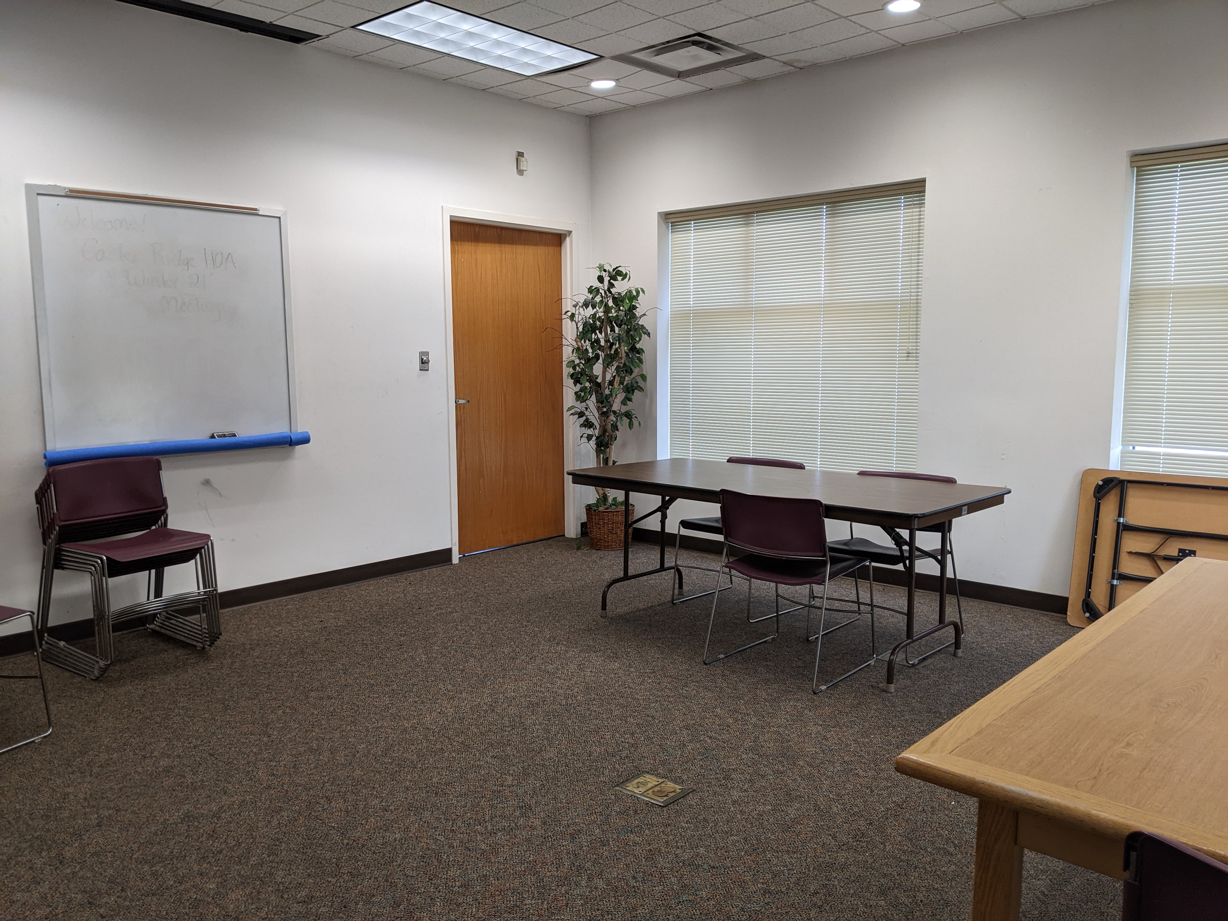 Photo shows a carpeted room, windows with blinds, folding tables and stackable chairs. A dry-erase board is on one wall.