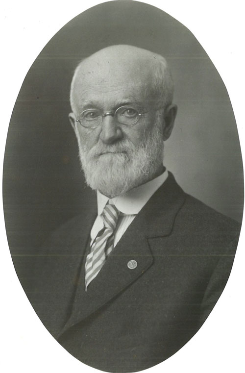 Black-and-white portrait of man in suit. The man is wearing glasses and is balding with white hair and a white mustache and beard.