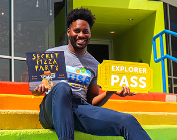 young man with Muse Knoxville t-shirt holds Secret Pizza Party book and large Explorer Pass golden ticket