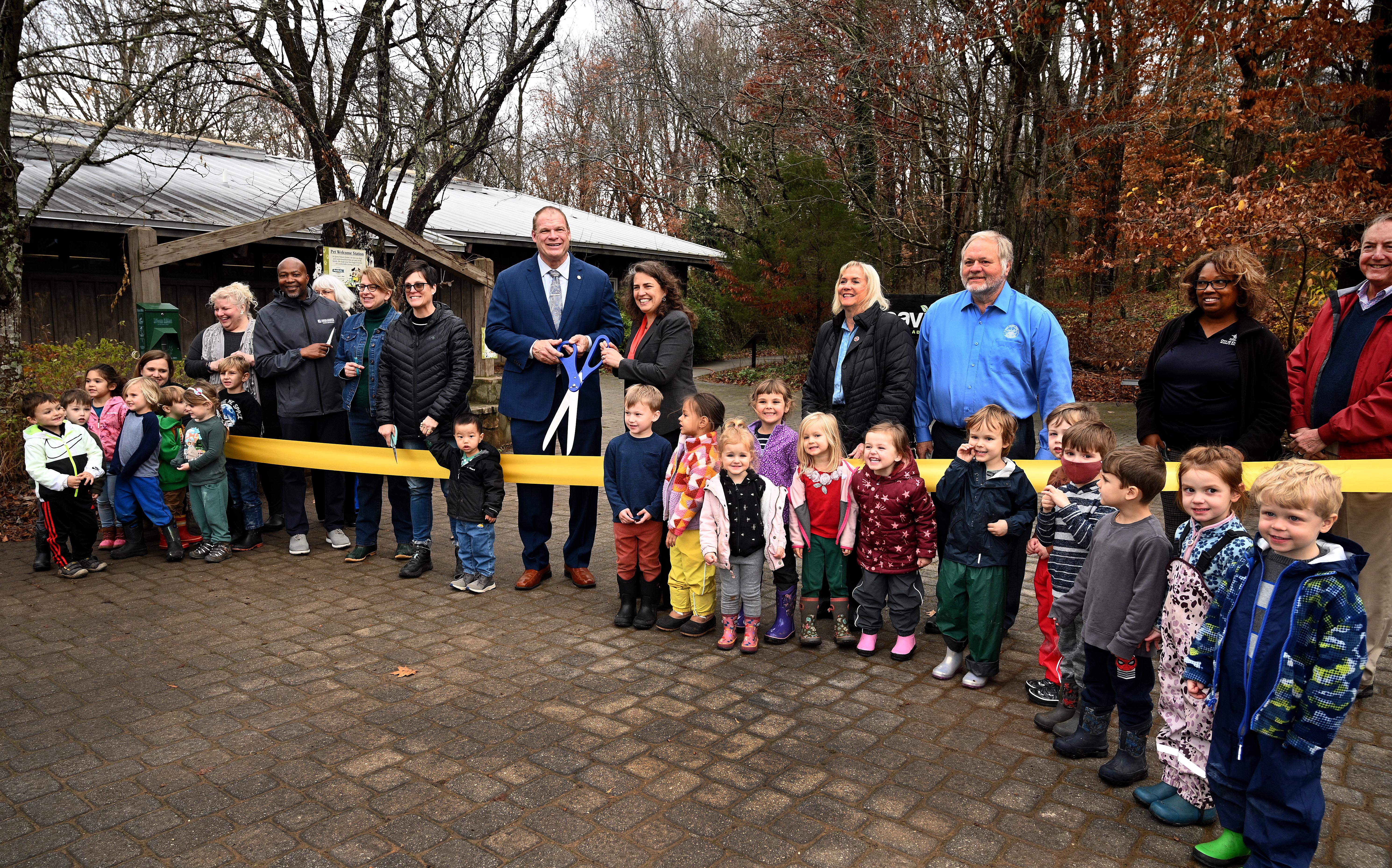 more than a dozen preschool children stand in front of large yellow ribbon with dignitaries behind holding scissors
