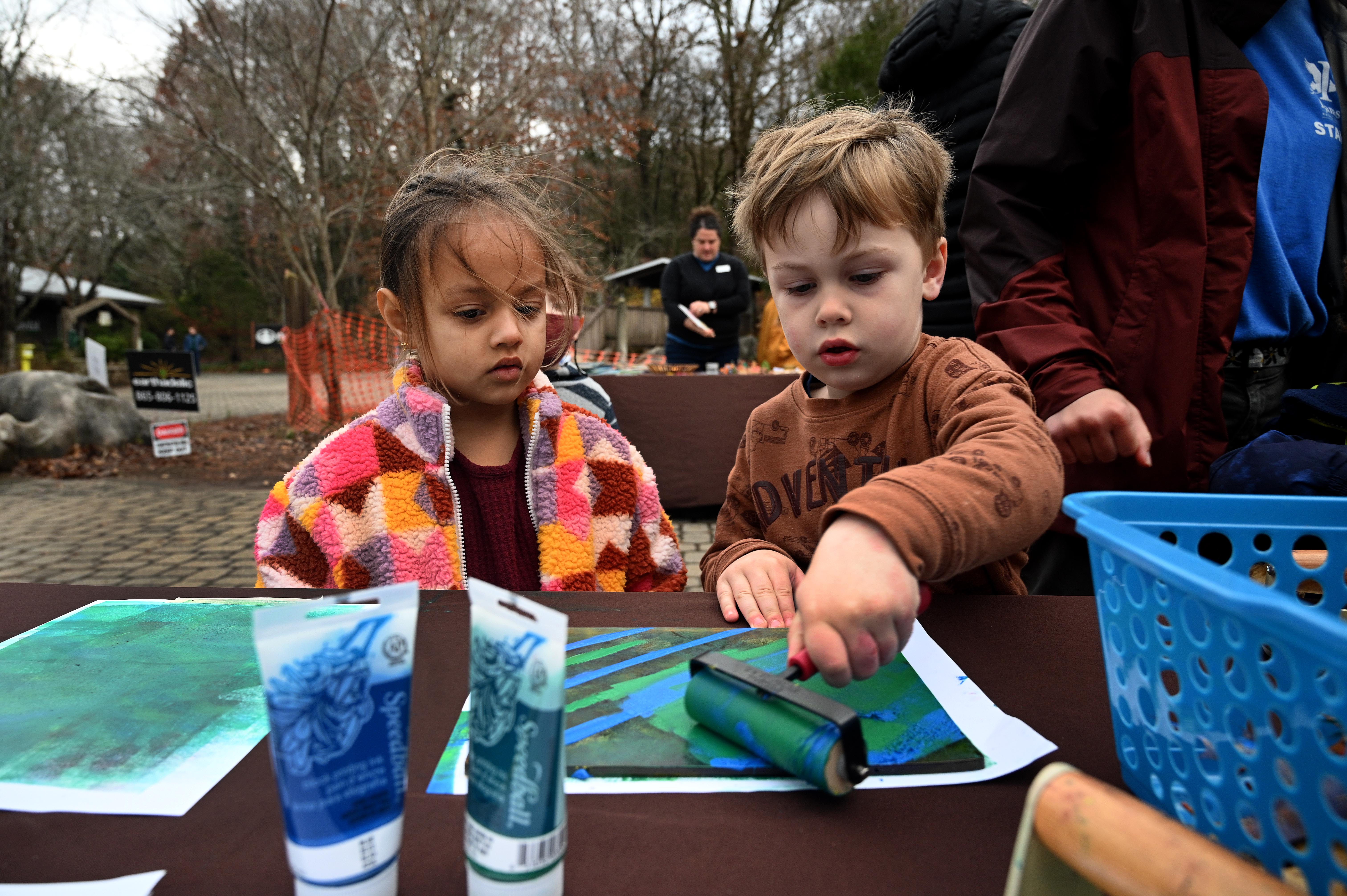 preschool boy rolls blue and green ink onto paper while preschool girl looks on