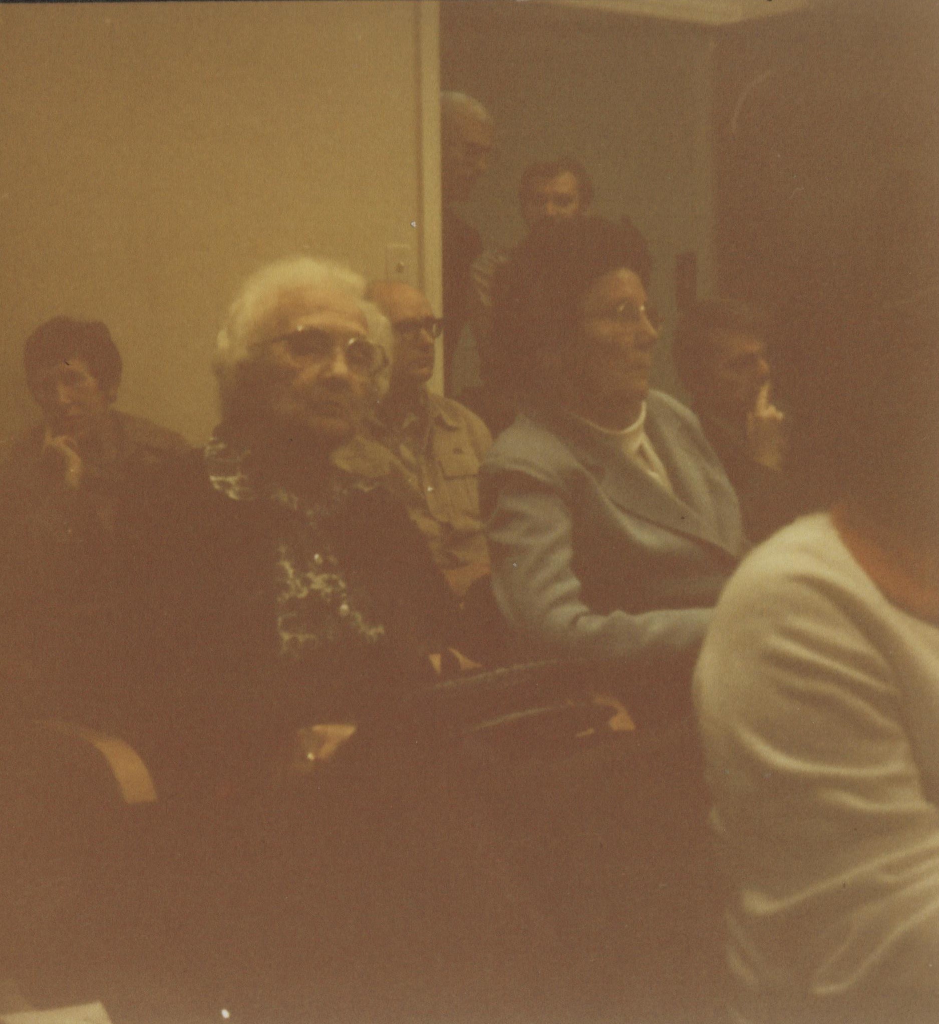 Color photograph of women seated in a room with a woman with white hair in middle of photo
