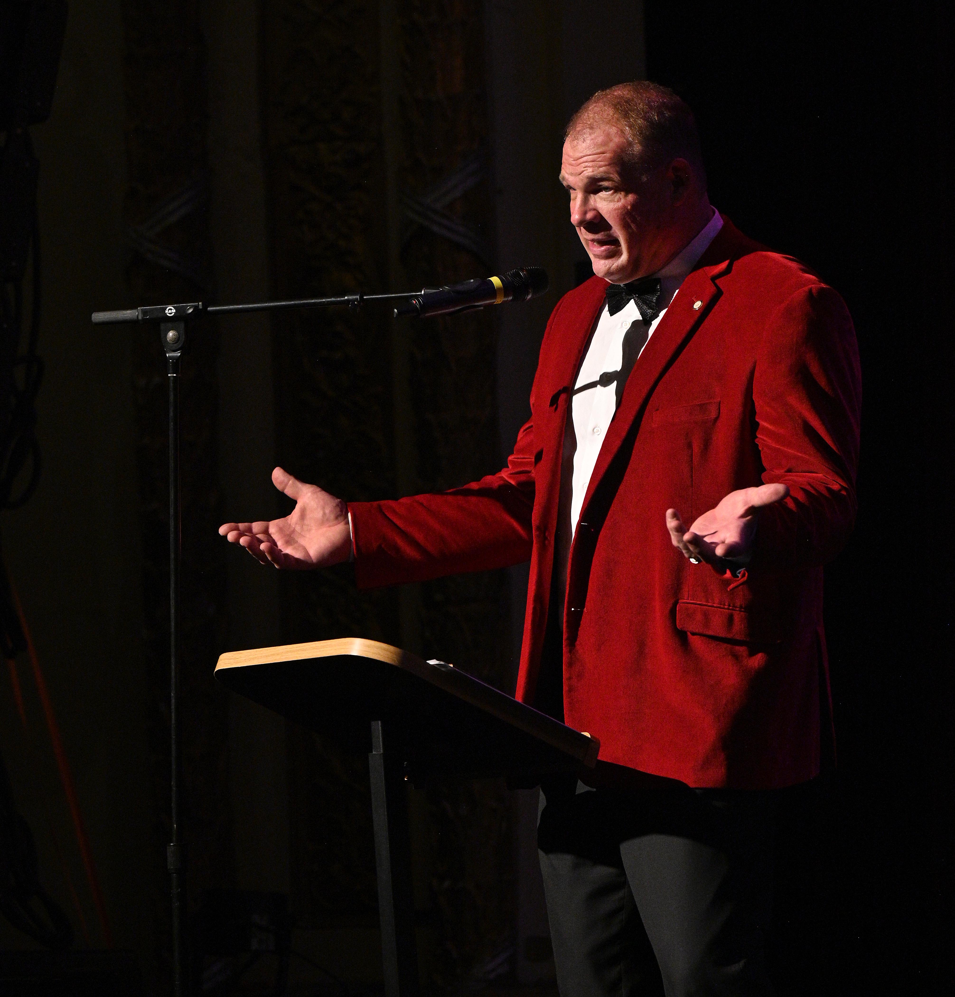 Mayor Glenn Jacobs wears a red velvet jacket, speaking at a microphone on a dark stage, standing with arms out, palms up