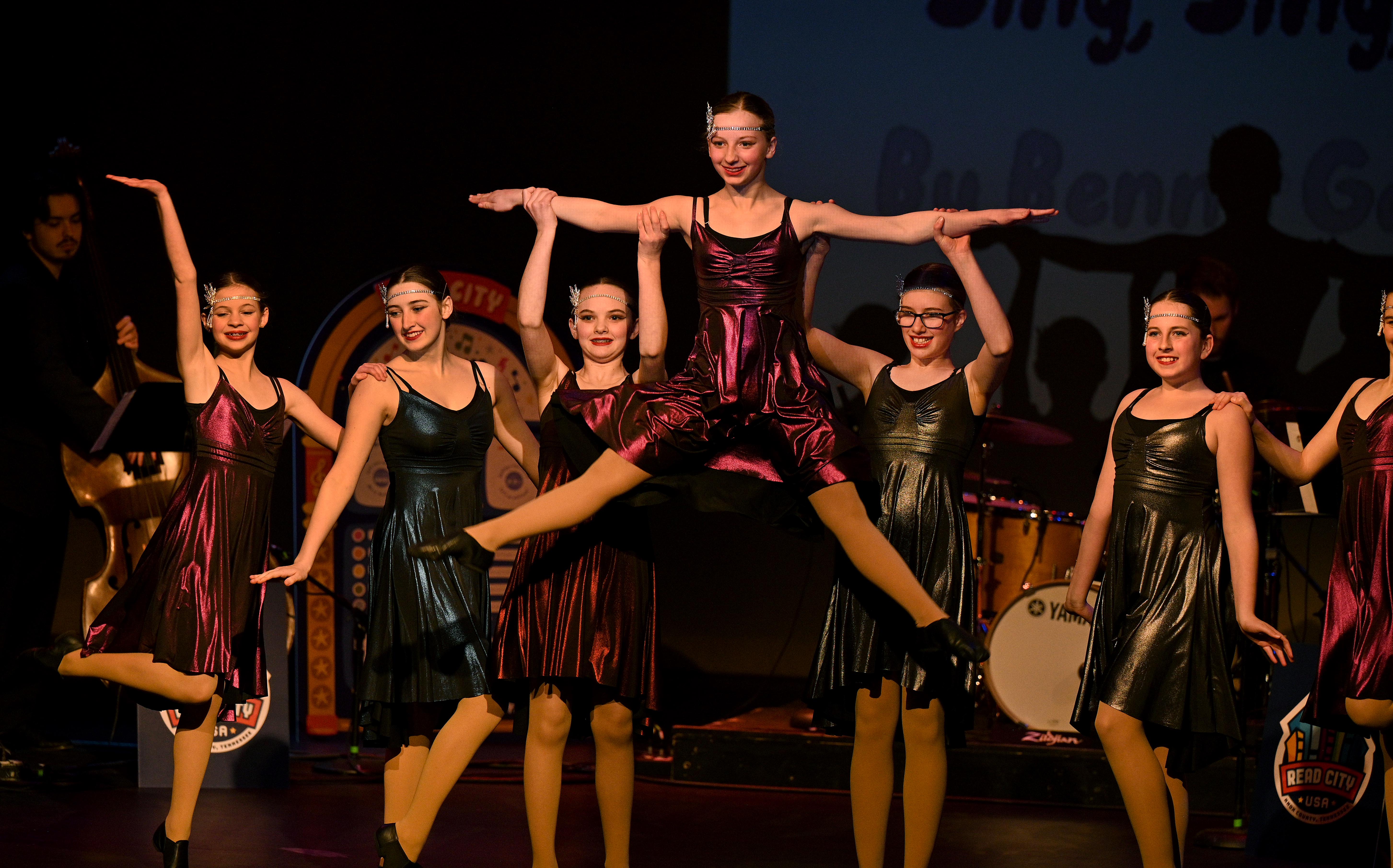 seven young dancers are arranged in a row on stage, mid-Charleston-style dance moves. Two in the middle hold one dancer aloft by her arms as she does the splits