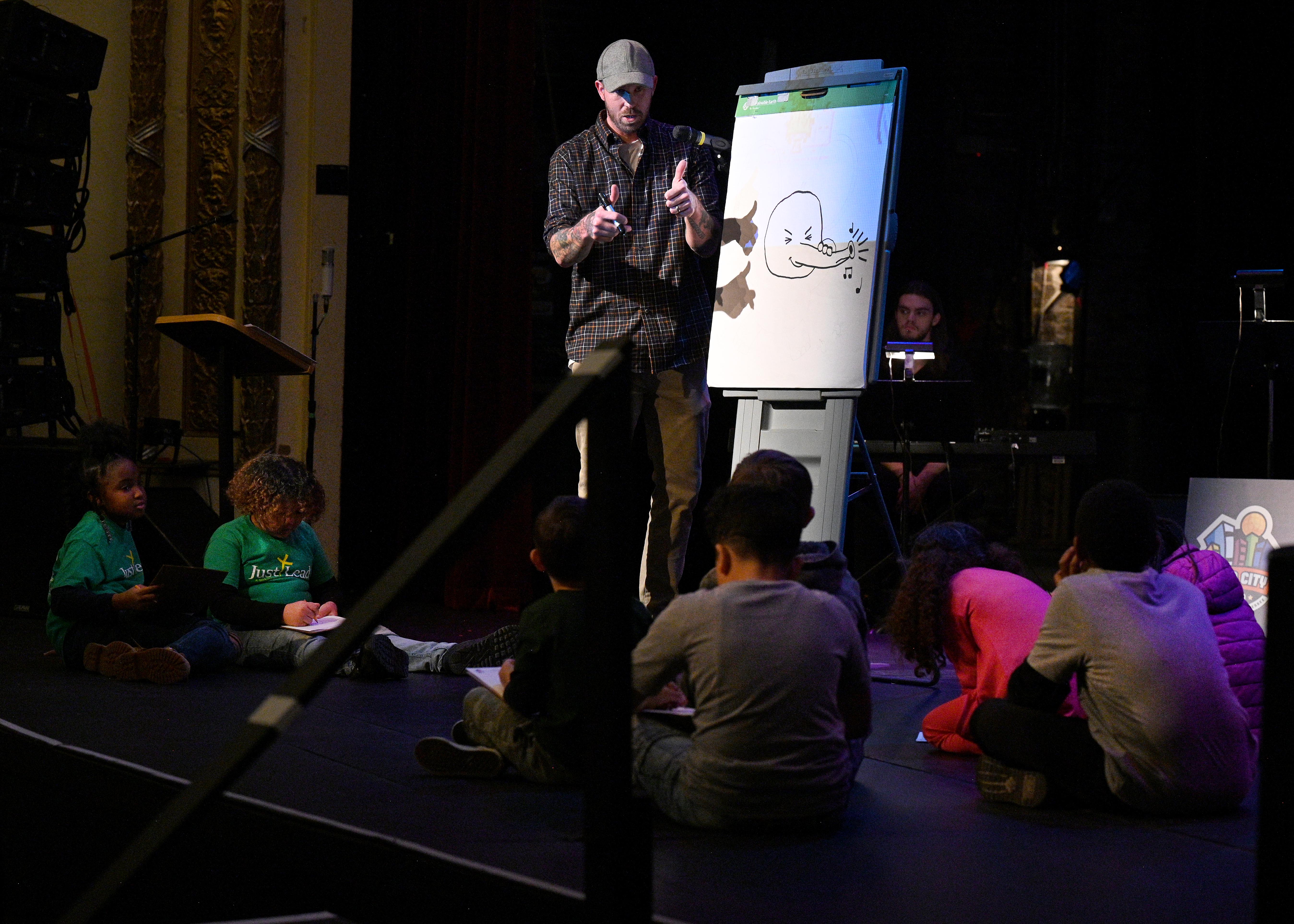 illustrator Daniel Wiseman stands at an easel on stage; a group of young children watch, seated