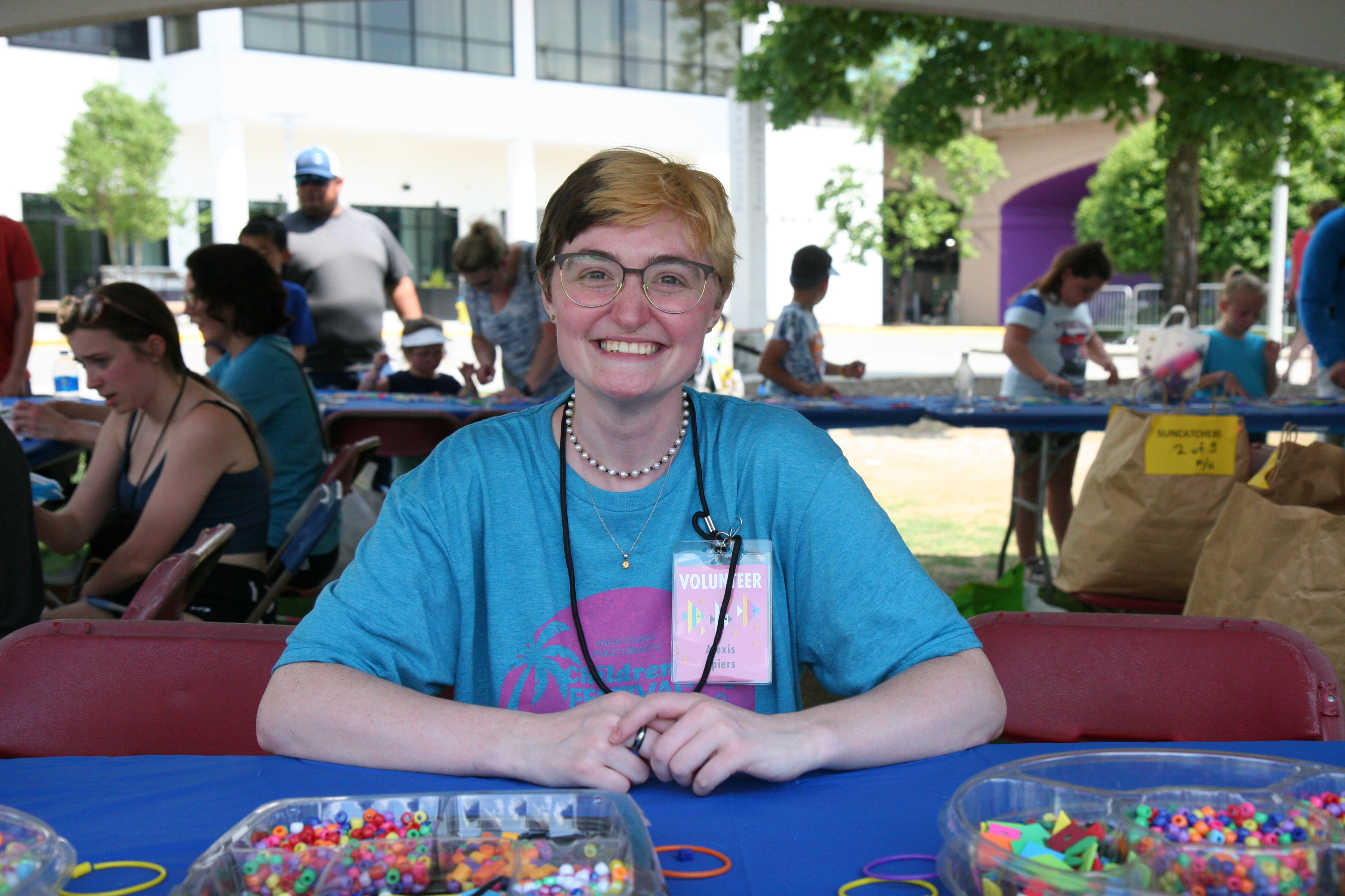 young woman seated at a table outdoors, smiling - festival volunteer