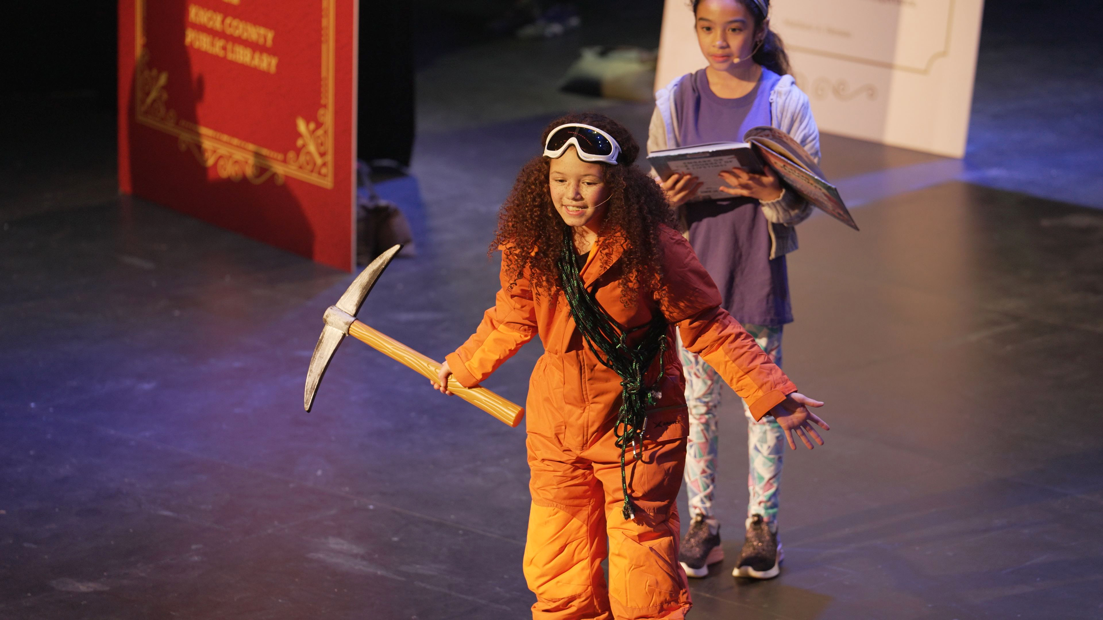 young girl dressed as mountain climber in orange snowsuit and pickaxe