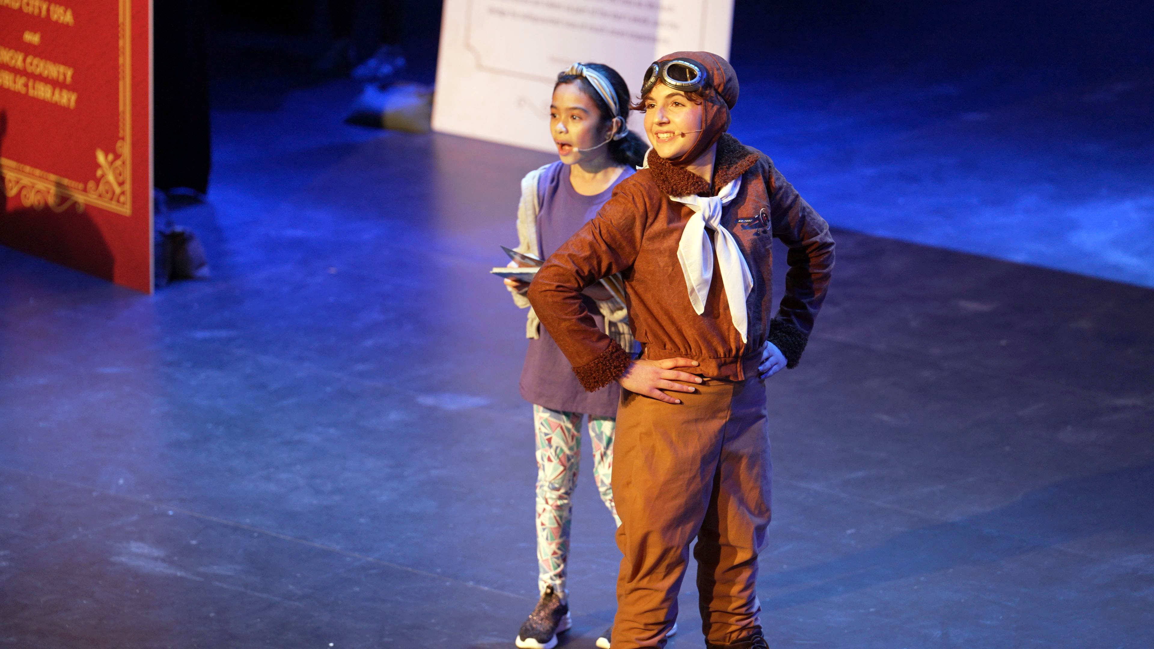 young woman dressed as Amelia Earhart in aviator hat, goggles, scarf, pilot jacket, riding pants