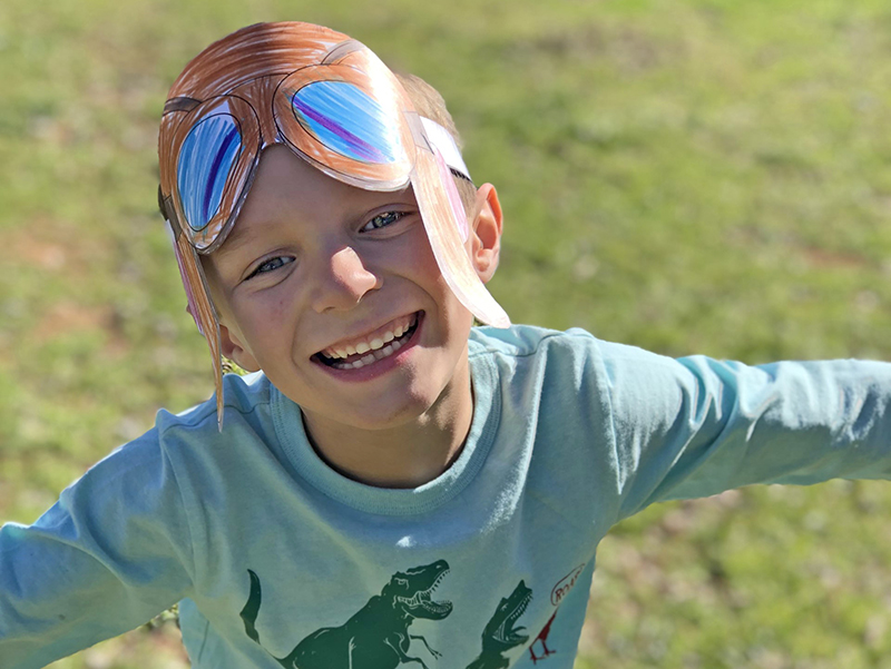 photo of young boy in paper aviator helmet, arms out like he's flying