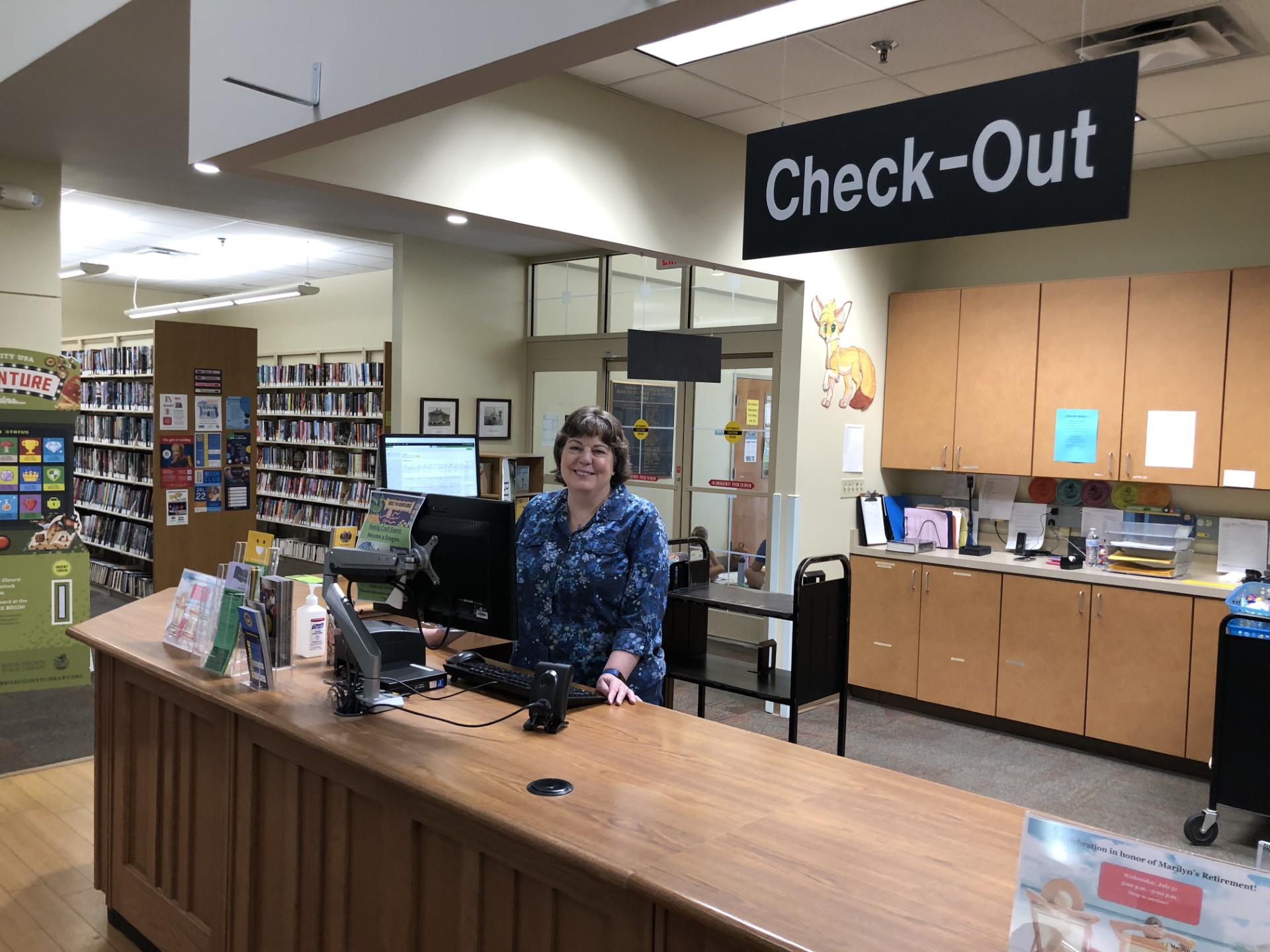 smiling woman at a library counter under a Check-Out sign