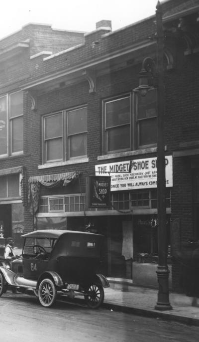 Black-and-white image of the exterior of a shop with a sign "The MIdget Shoe Shop" above the door.  The sign is partially obscured by a lamp post. A 1920s automobile is parked on the street in front of the shop. 