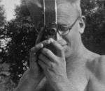 A spectacled man holds a home movie camera to his eye