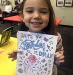 young girl holds a coloring sheet with cupcake and the words Happy Birthday Dolly
