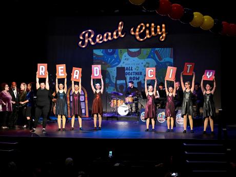 Young dancers on a stage hold cards that spell R-E-A-D C-I-T-Y with Read City in script marquee letters behind them