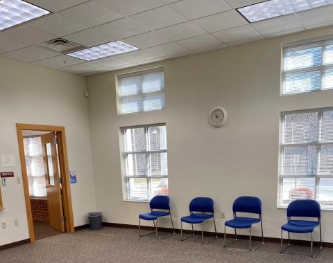 Photo shows carpeted room with windows with blinds. Along the wall, four stackable chairs with plastic seat and back.