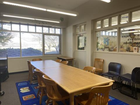 Photo shows a carpeted room with windows to the outside and into the library main room, wooden tables and chairs, and stackable chairs with plastic seat and back.