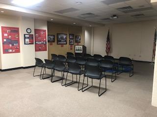 Photo shows carpeted room with flags, lectern, three rows of five chairs each. Stackable chairs have plastic seat and back.