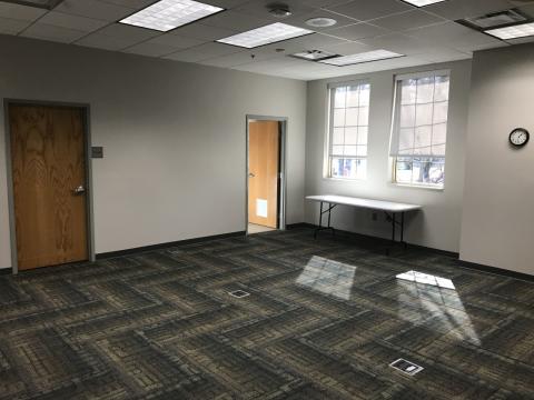 Photo shows carpeted room with table underneath windows; power outlets in middle of floor
