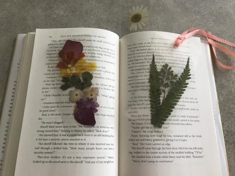 Staff have created a book mark example with pressed flowers that have been laminated. Book mark is laying on top of a book.