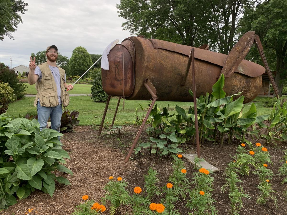 Entomologist standing outdoors near plants and statue of bug.