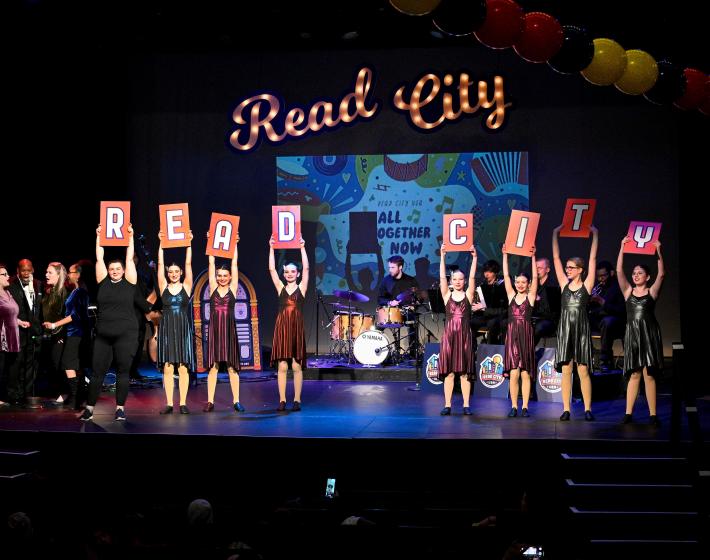Young dancers on a stage hold cards that spell R-E-A-D C-I-T-Y with Read City in script marquee letters behind them