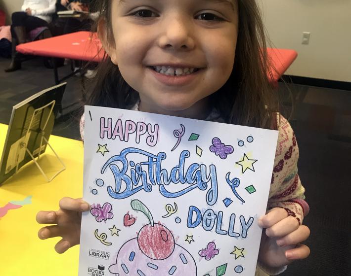 young girl holds a coloring sheet with cupcake and the words Happy Birthday Dolly